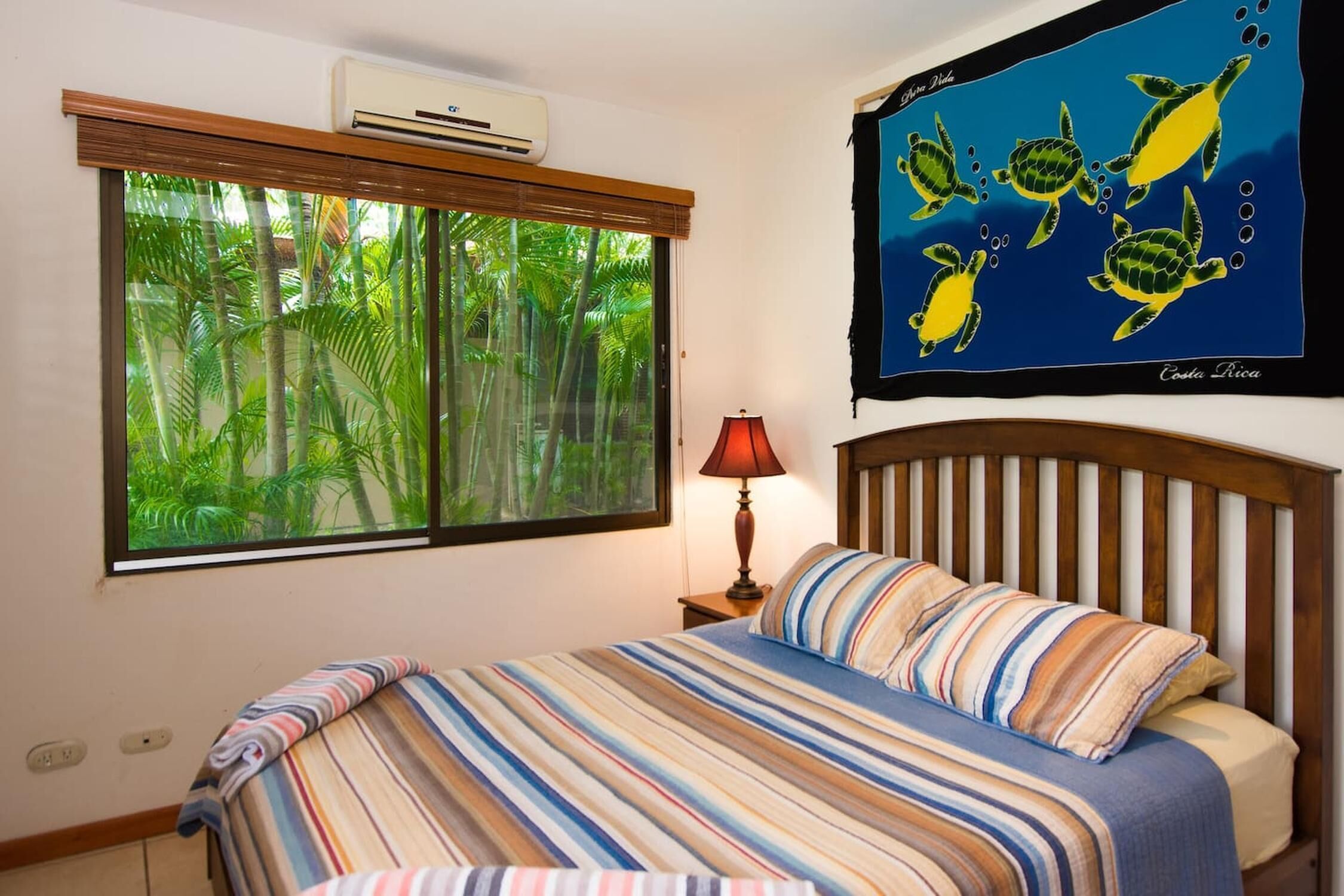 Un dormitorio con un ventanal grande, una cama con ropa de cama a rayas, una mesita de noche con lámpara y un cuadro de pared con tortugas marinas.