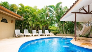 Une piscine entourée de palmiers, avec des chaises longues blanches et un hamac.