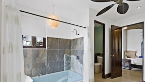 Une salle de bain moderne avec une douche carrelée en pierre, une baignoire aux carreaux de mosaïque bleus, un ventilateur de plafond et une porte en bois donnant sur une autre pièce.