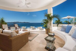 A sunlit outdoor lounge area with a curved white sofa, wicker furniture, and a view of the sea. - Exquisite Hilltop Villa with Luxurious Modern Design, Sweeping Ocean Views & Sparkling Infinity Pool (Cabo Velas)