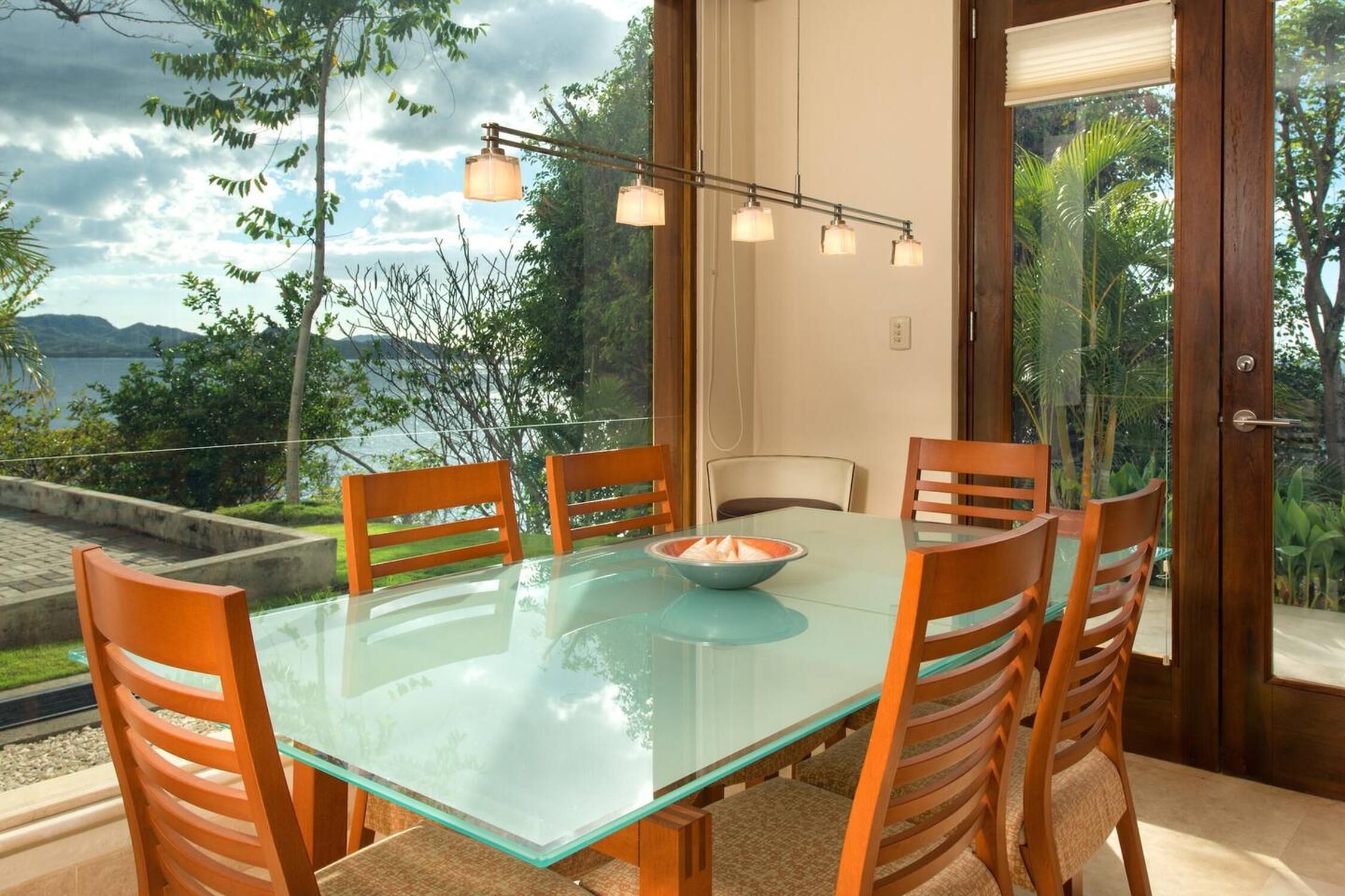 A dining area with a glass table and wooden chairs, overlooking a scenic view of a lake and mountains.