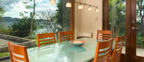 A dining area with a glass table and wooden chairs, overlooking a scenic view of a lake and mountains.