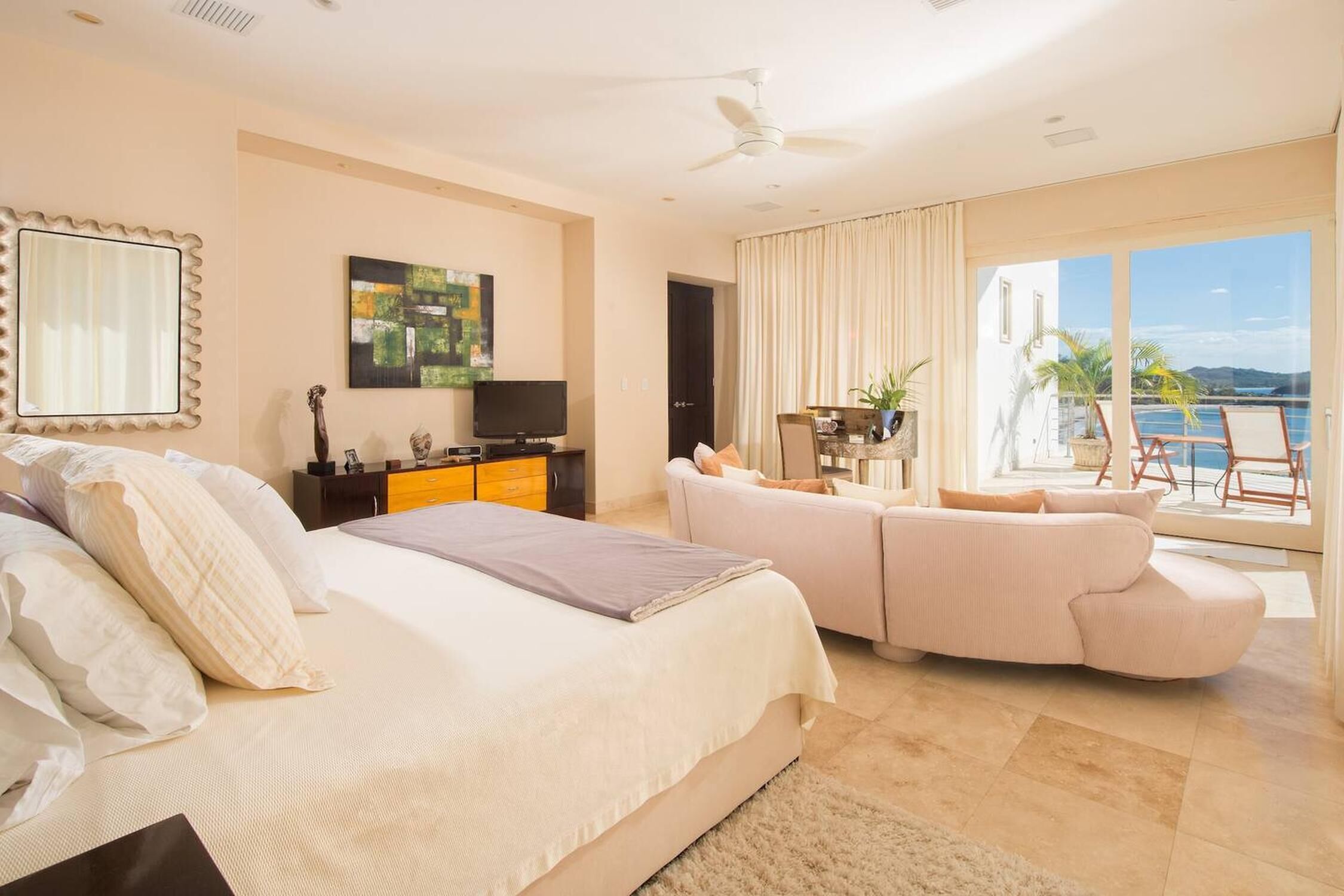 A hotel room with a bed, a sofa, a TV, a ceiling fan, and a balcony with a view of the sea.