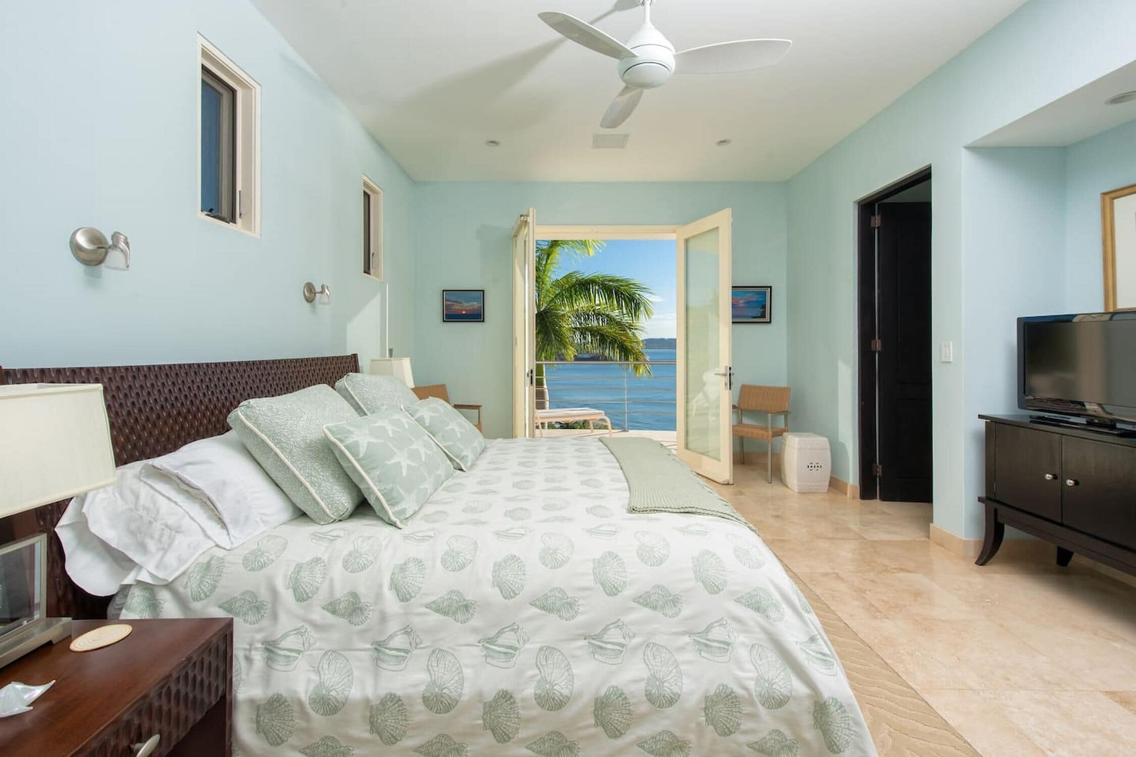 A bedroom with a bed, a TV, a ceiling fan, a window with a view of the sea, and a door leading to another room.