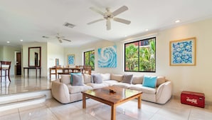 A spacious living area with a sofa, coffee table, and dining set. There are ceiling fans, large windows with views of greenery, and artwork on the walls. - Recently Renovated Luxury Condo with Resort-Style Beach Club Access and Ocean Views (Cabo Velas)
