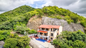 A two-story building with a red roof, a swimming pool, and a view of a lush green hillside. - Coastal-Inspired Condo w/ Infinity Pool & Oceanview Terrace (Cabo Velas)