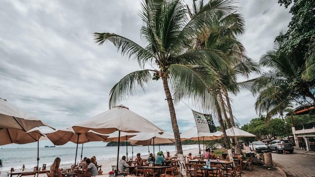 A beachside restaurant with wooden tables and chairs, umbrellas, and a palm tree. - Coastal-Inspired Condo w/ Infinity Pool & Oceanview Terrace (Cabo Velas)