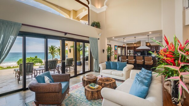 A spacious living area with a large sliding glass door leading to a balcony overlooking the ocean. There are wicker chairs and a white sofa with blue cushions. A kitchen area is visible in the background with wooden bar stools. - Stunning Luxury Mansion in Playa Flamingo with Six Bedrooms and Beachfront Access (Cabo Velas)