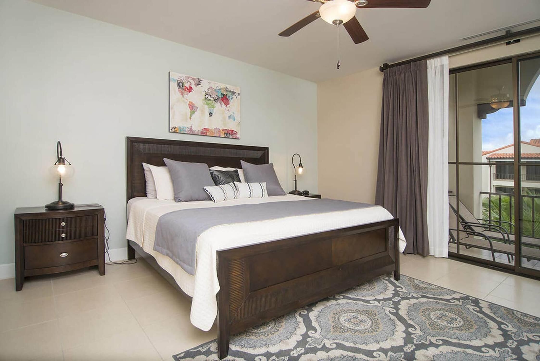 A bedroom with a large bed, two bedside tables, a ceiling fan, a world map artwork, and a view of a balcony.