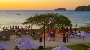 Un lugar frente a la playa con mesas y sillas al aire libre, sombrillas y sillones reclinables.
