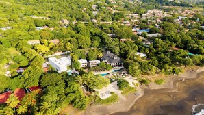 Vista aérea de un pueblo costero con edificios residenciales, una playa y una colina con vegetación.