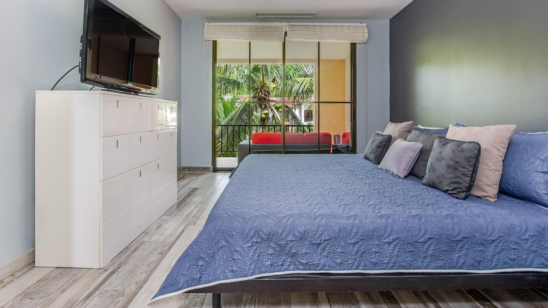 Une chambre moderne avec un grand lit, un téléviseur posé sur un meuble et un balcon offrant une vue sur les palmiers.
