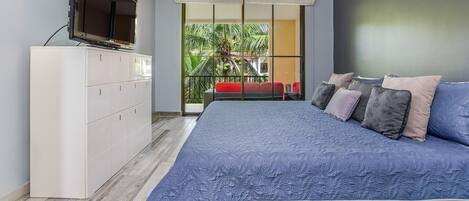 Une chambre moderne avec un grand lit, un téléviseur posé sur un meuble et un balcon offrant une vue sur les palmiers.