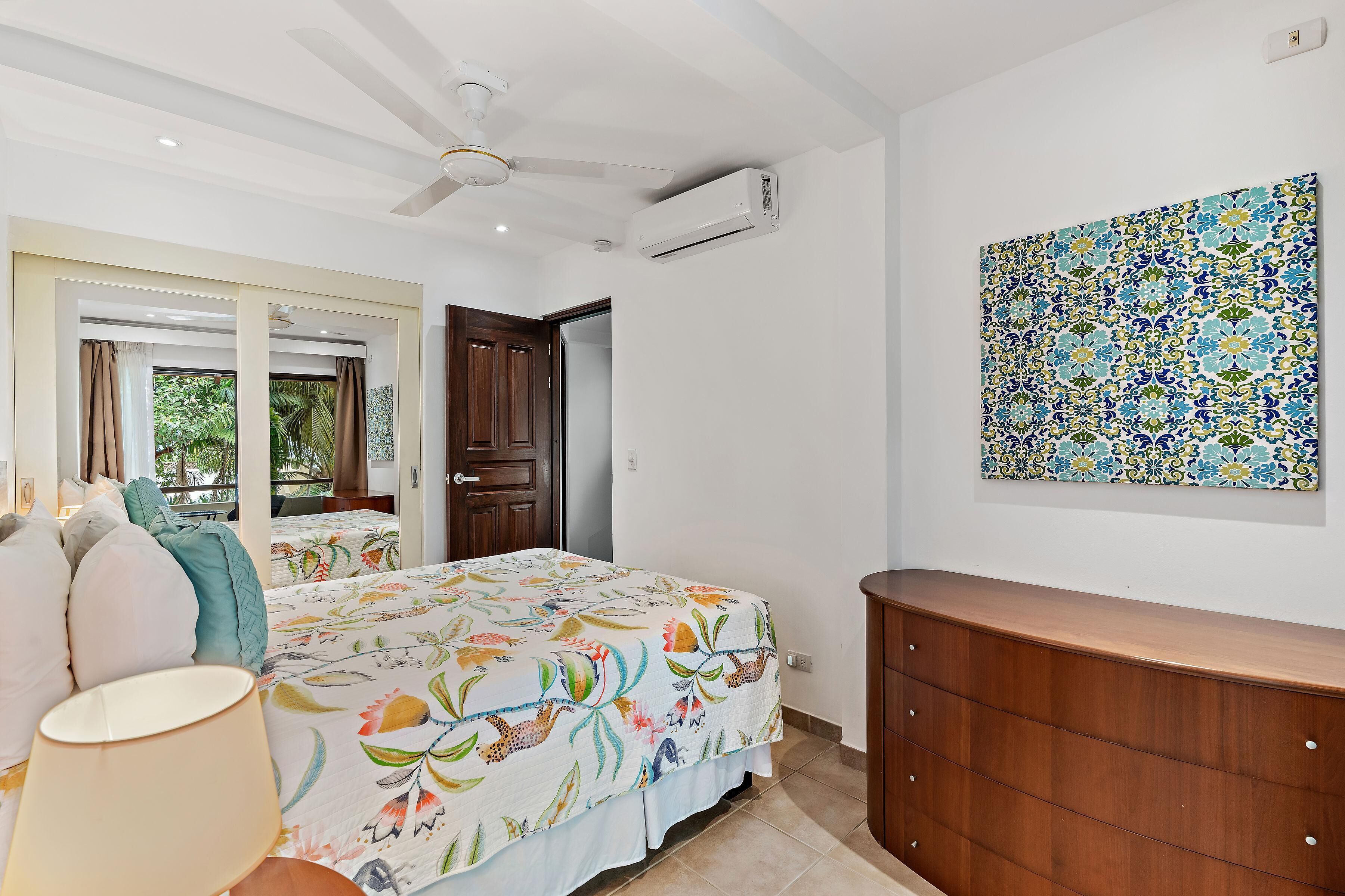 A bedroom with a bed, a wooden dresser, a ceiling fan, an air conditioning unit, a window with curtains, and a decorative wall piece.