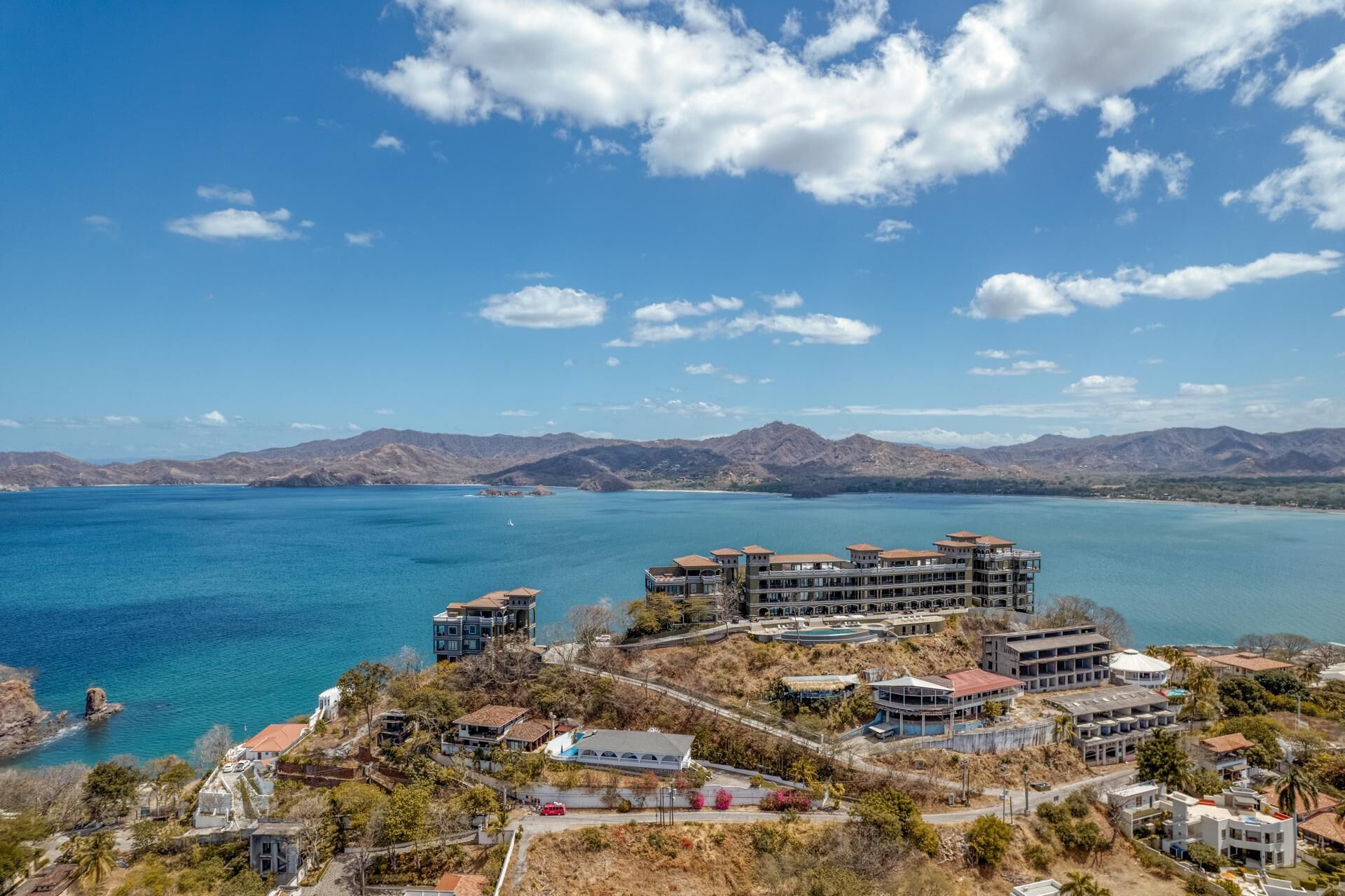 A coastal resort with multiple buildings, a pool, and a view of the sea and mountains.