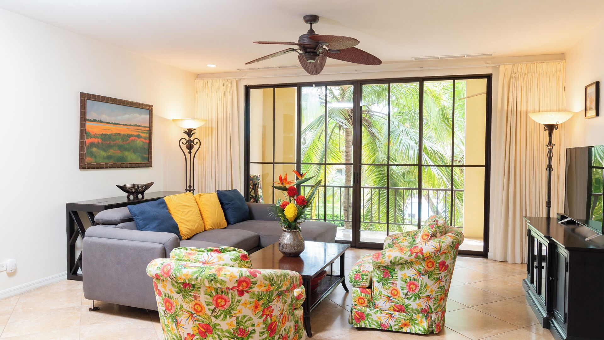 A living room with a sofa, two floral armchairs, a coffee table, a painting, a ceiling fan, and a large window with curtains.