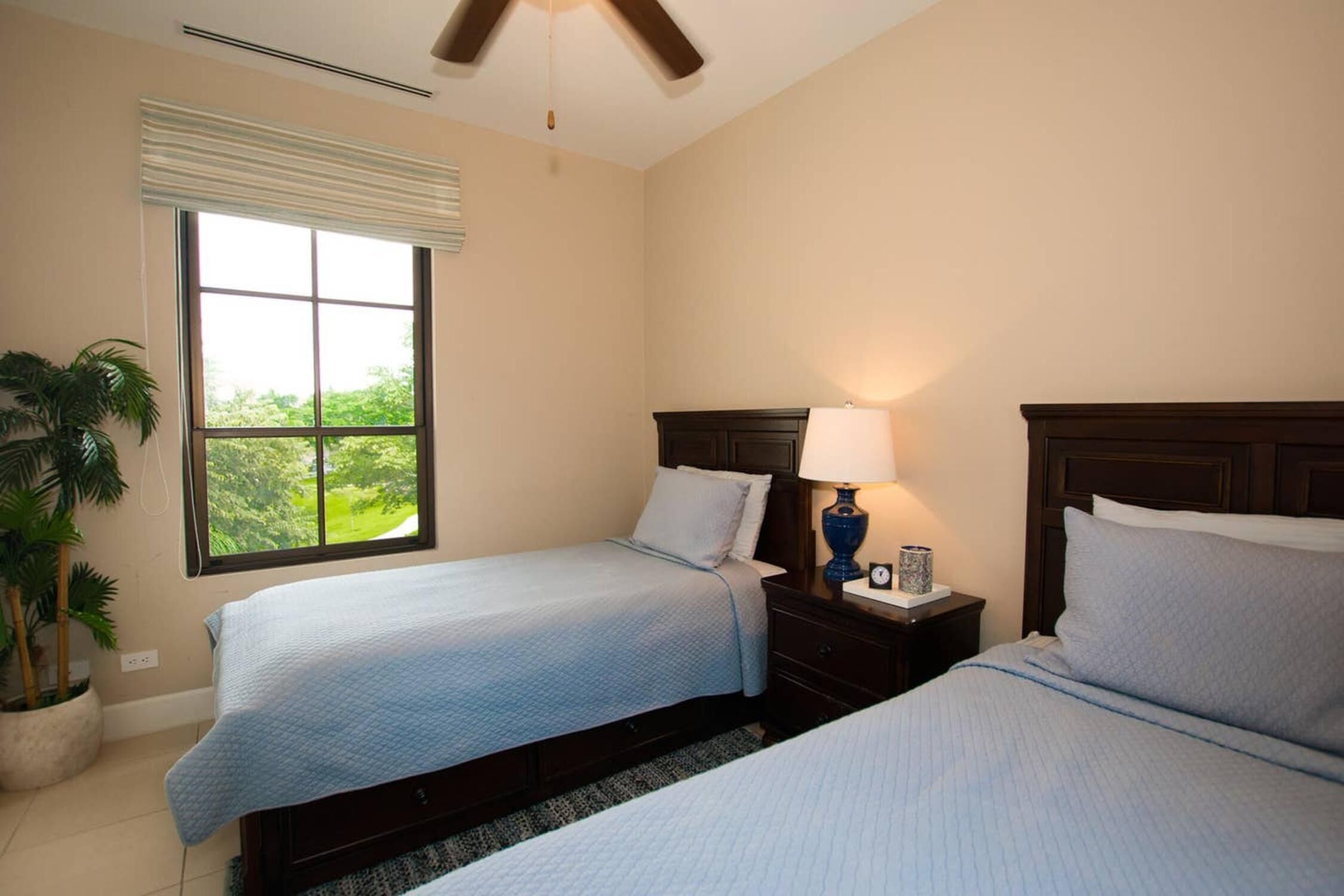 A hotel room with two beds, a large window with blinds, a ceiling fan, and a nightstand with a lamp.