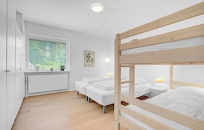 Une chambre avec des lits superposés, une fenêtre avec des stores, un tableau encadré et une table de chevet avec une lampe.