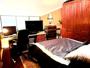Comfortable room with a bed, desk, and wardrobe, featuring a window with blinds and a vase with flowers.