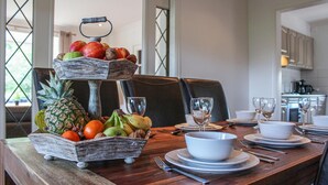 Une table à manger dressée avec des assiettes, des bols et des couverts, une présentation de fruits et une vue sur la cuisine.