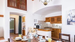 A dining area with a wooden table set for breakfast, a kitchen in the background, and a view of the street. - Villa Jacaranda V3 by Interhome (Albufeira)