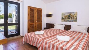 A hotel room with two beds, a sliding glass door to the outside, a wooden door, a painting on the wall, and a nightstand with a lamp. - Villa Jacaranda V3 by Interhome (Albufeira)