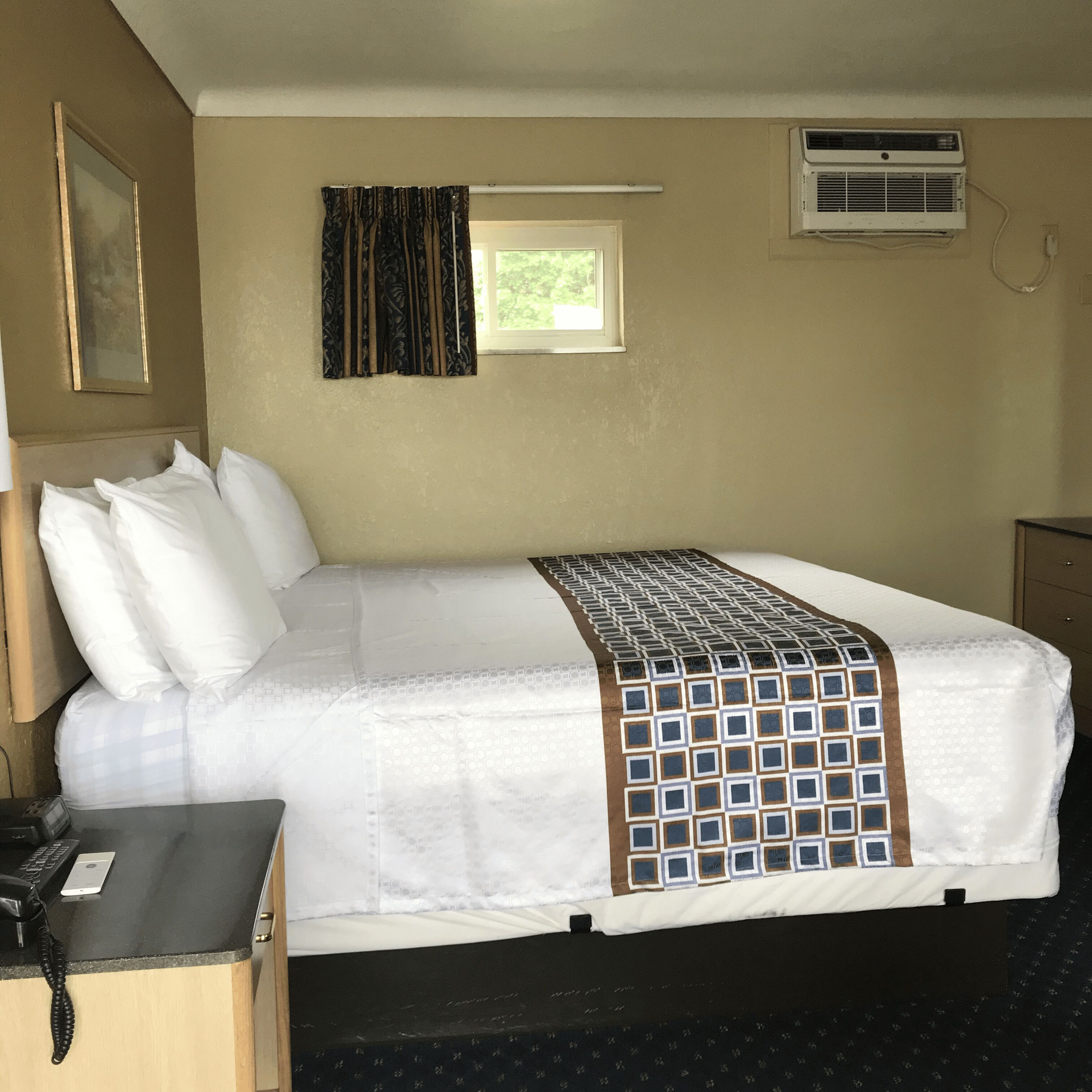 A hotel room with a bed, a desk with a telephone, a window with curtains, and an air conditioning unit.