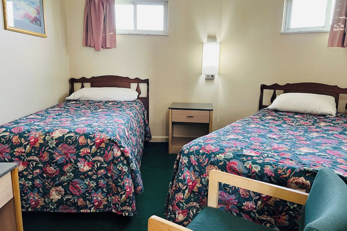A room with two beds, each with floral bedding, a bedside table, a lamp, and a window.