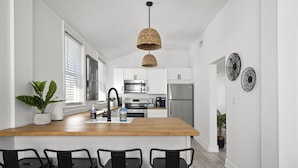 A modern kitchen with a wooden island, black bar stools, stainless steel appliances, and decorative wall art. - Surf Park House – Historic Jax Beach Home, 3 Blocks to Beach, Pet Friendly (Jacksonville Beach)