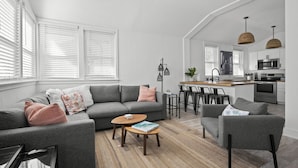 A modern living room with a gray sofa, a coffee table, and a kitchen area with a bar and seating. - Surf Park House – Historic Jax Beach Home, 3 Blocks to Beach, Pet Friendly (Jacksonville Beach)