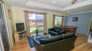 A living room with a black leather sofa, a coffee table, a TV on a wooden stand, and a dining area with a table and chairs. - McLeods • Pet-Friendly Central Beach House in Agnes Water • Walk Everywhere (Agnes Water)