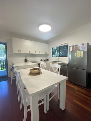 A compact kitchen with white cabinets, a dining table with chairs, a refrigerator, and a window with a view of greenery.