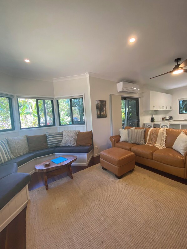 A spacious living area with a sofa, ottoman, and coffee table, large windows with a view of greenery, and a kitchen area with cabinets and a TV.