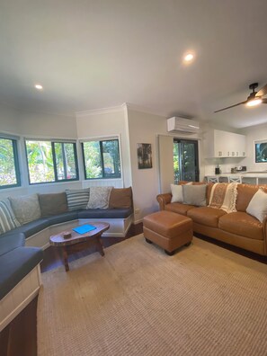 A spacious living area with a sofa, ottoman, and coffee table, large windows with a view of greenery, and a kitchen area with cabinets and a TV. - Merindah - Pet-Friendly, Coastal Living at its Best (Agnes Water)