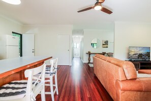 A modern kitchen with a wooden countertop and white chairs, a living area with a leather sofa and a flat-screen TV, and a hallway leading to a door.