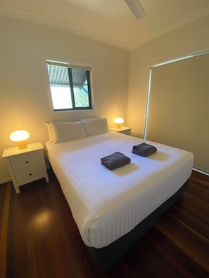 Habitación con cama individual, ventana, dos mesitas de noche con lámparas y una cama bien hecha con dos toallas dobladas.