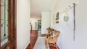 A hallway with a wooden floor, a wicker chair, a dining table with chairs, and a coat rack with bags.