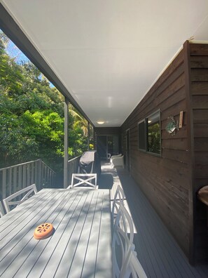 A covered outdoor deck with a table and chairs, a grill, and a view of the surrounding greenery.