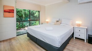 A modern bedroom with a large bed, two bedside tables, a window with a view of greenery, and an air conditioning unit.