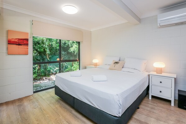 A modern bedroom with a large bed, two bedside tables, a window with a view of greenery, and an air conditioning unit.