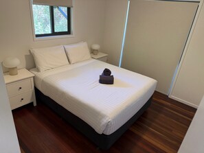 A neatly made bed with a folded towel, a bedside lamp, and a window with a view of trees.