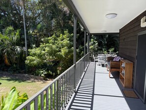 A covered outdoor deck with a railing, a seating area with a table and chairs, and a view of dense tropical vegetation.