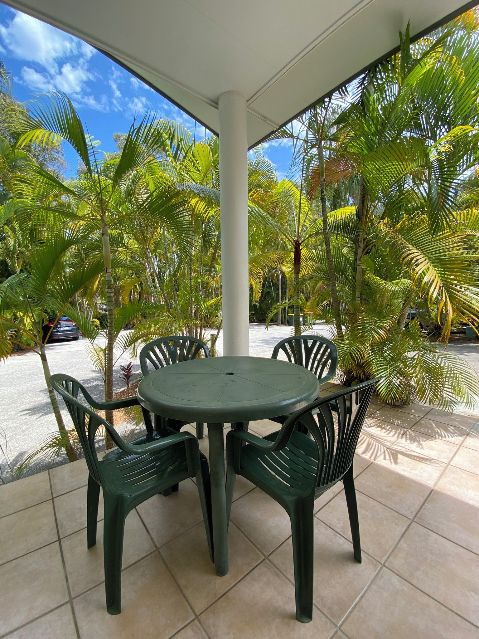 A covered outdoor seating area with a round table and four chairs, surrounded by palm trees and a clear sky.