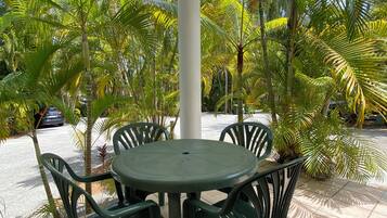 A covered outdoor seating area with a round table and four chairs, surrounded by palm trees and a clear sky.