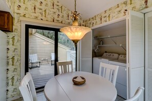 Une pièce avec une table ronde, des chaises blanches, un luminaire suspendu, une porte donnant sur l’extérieur et un coin buanderie avec une machine à laver et des étagères.
