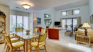 A spacious living room with a glass dining table, comfortable seating, a television, and a balcony with a view.