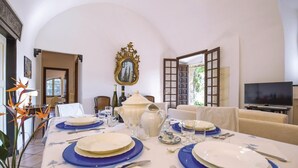 Un coin repas avec une table dressée pour quatre personnes, un téléviseur et une vue sur un jardin par une porte ouverte.