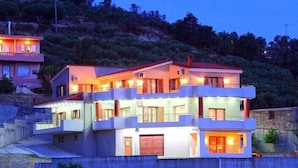 A multi-story building with balconies and a red roof, surrounded by hills and trees. - Villas metoxi (Heraklion)