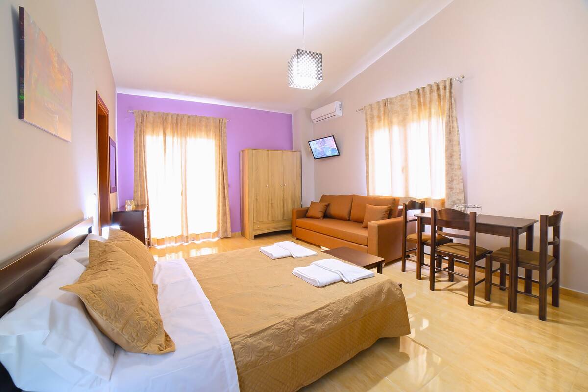 A hotel room with a large bed, a sofa, a dining table with chairs, and a TV on the wall.