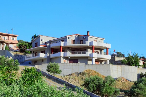A multi-story residential building with balconies, situated on a hillside with greenery in the foreground. - Villas metoxi (Heraklion)